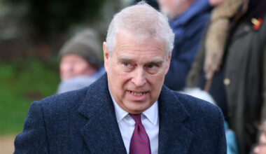 Prince Andrew has white gray hair and wears a large overcoat with a white shirt and red tie underneath