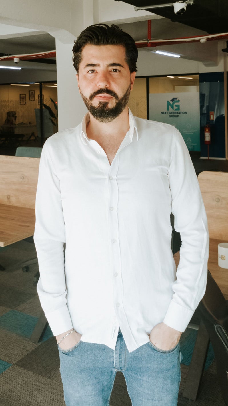 A man with a beard stands confidently in an office environment, wearing a white button-up shirt and blue jeans, with wooden tables and a modern office backdrop.