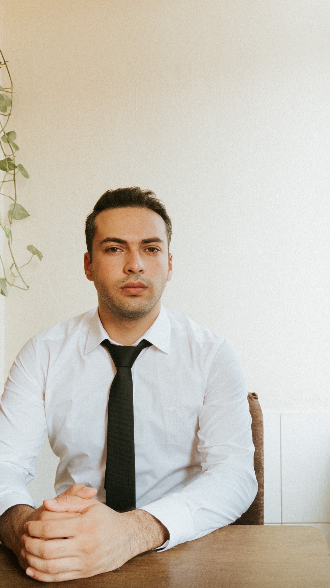 A man in a white shirt and black tie sits at a wooden table, with his hands clasped together, staring directly at the camera. The background features a light-colored wall with a trailing plant.