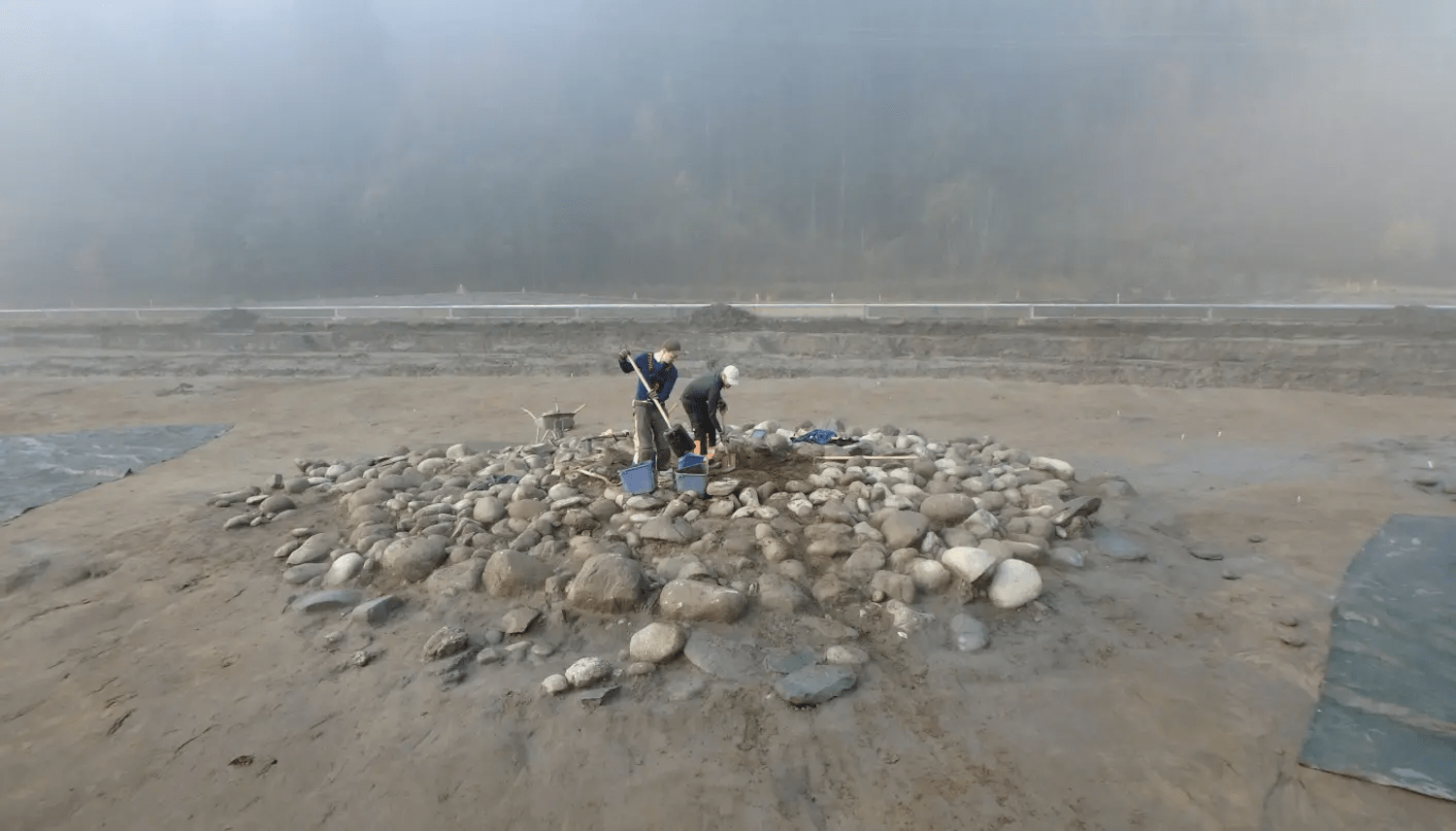 Two archaeologists digging among burial cairn stones on foggy day
