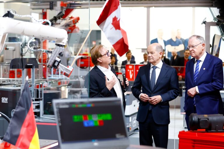 German Chancellor Olaf Scholz talking with two other men in suits at an industrial fair booth with a Canadian flag in the background.