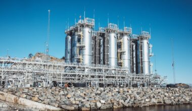 Tall cylindrical storage tanks situated on a stone jetty in industrial coastal location with walkways and other structures arrayed around the top and sides of the tanks