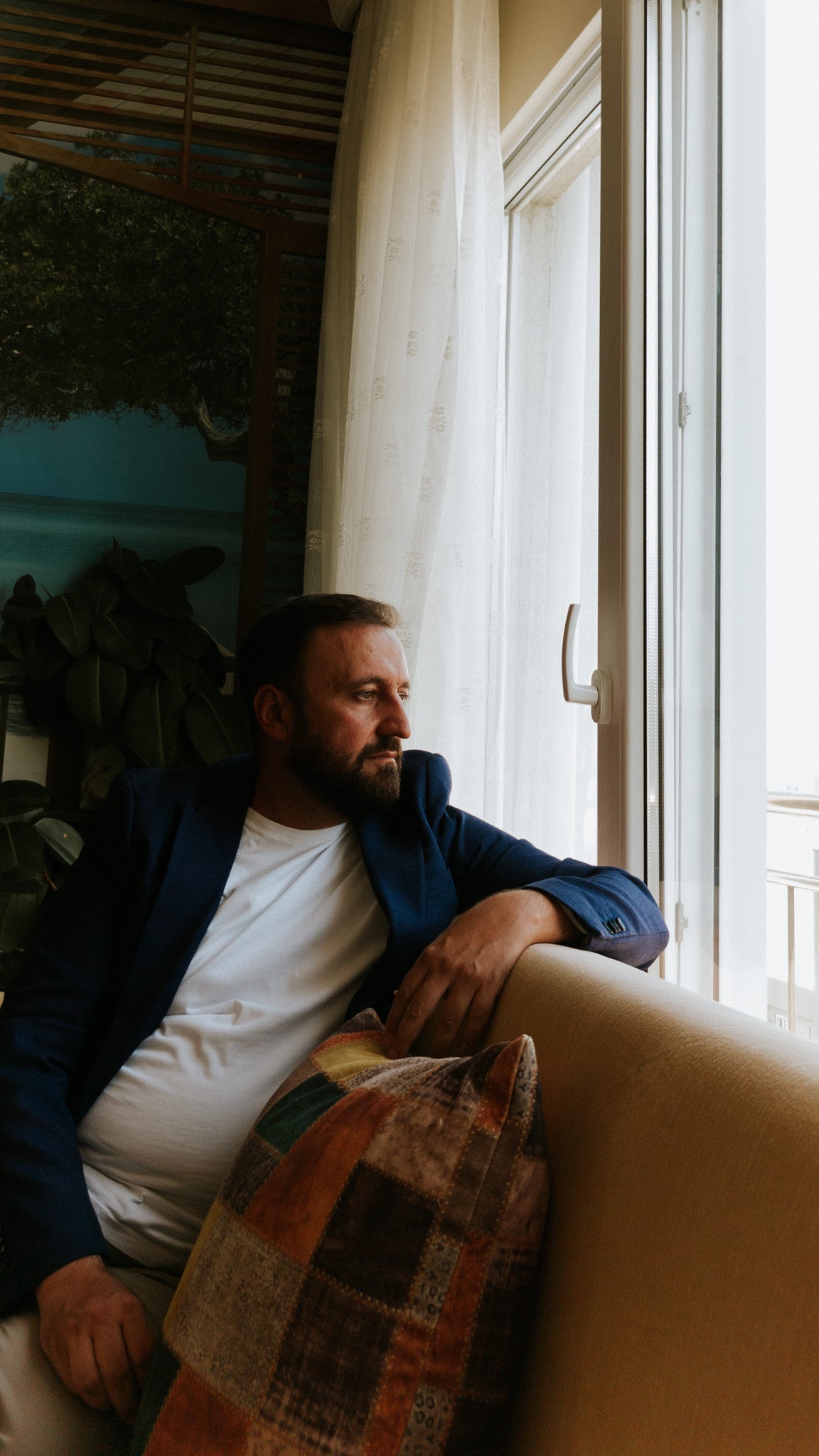 A man with a beard and short haircut, dressed in a blazer and white shirt, sits on a sofa looking contemplatively out of a window with sheer curtains. In the background, a large painting of a calm sea and a tree is visible along with indoor plants.