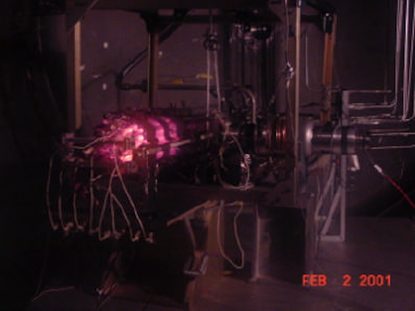 A laboratory setup features electronic equipment and wires, with a glowing pink light source in the center—resembling a miniature nuclear reactor; the date stamp reads 