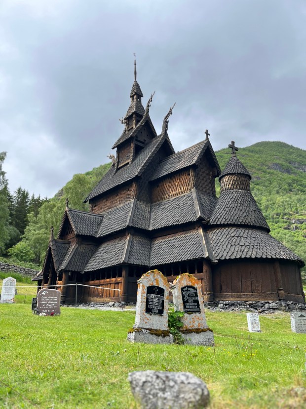 A dark gray Viking Stave church with a bright green mountain behind it
