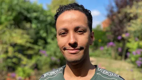 Aziz has short dark hair and is wearing a green T-shirt. He's standing in a garden with shrubbery and flowers behind him. It's a sunny day and the sky is blue.