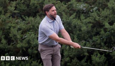 A man with brown hair and beard, wearing a light blue top an brown trousers swings a golf club with bushes behind him.