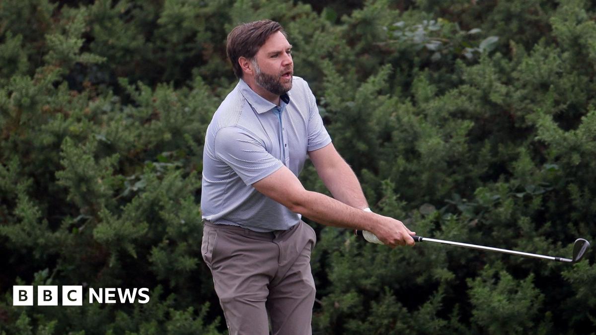 A man with brown hair and beard, wearing a light blue top an brown trousers swings a golf club with bushes behind him.