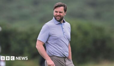 JD Vance looking slightly off camera. He is on a golf course. He is only visible from the waist up. He is wearing a light-coloured polo shirt with a thin horizontal pinstripe design. He is wearing dark beige trousers. He has dark hair and a dark beard. He is holding a golf club in his right hand, which is just out of shot and has his left hand in his pocket.