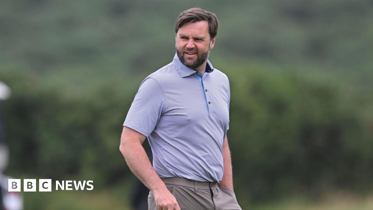JD Vance looking slightly off camera. He is on a golf course. He is only visible from the waist up. He is wearing a light-coloured polo shirt with a thin horizontal pinstripe design. He is wearing dark beige trousers. He has dark hair and a dark beard. He is holding a golf club in his right hand, which is just out of shot and has his left hand in his pocket.
