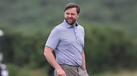 PA Media JD Vance looking slightly off camera. He is on a golf course. He is only visible from the waist up. He is wearing a light-coloured polo shirt with a thin horizontal pinstripe design. He is wearing dark beige trousers. He has dark hair and a dark beard. He is holding a golf club in his right hand, which is just out of shot and has his left hand in his pocket.
