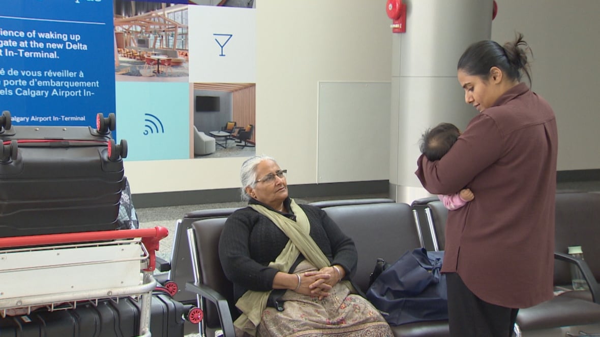 A woman stands holding her baby while an older woman sits on a chair watching them. They have a cart full of luggage beside them.