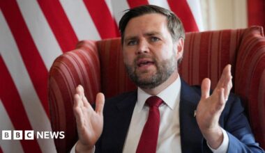 JD Vance sits in a chair, wearing a suit and tie. He has his mouth open and is speaking, with his hands in the arms to emphasise a point.