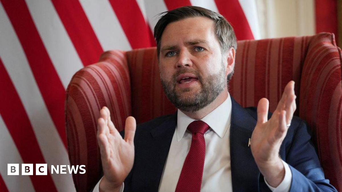 JD Vance sits in a chair, wearing a suit and tie. He has his mouth open and is speaking, with his hands in the arms to emphasise a point.
