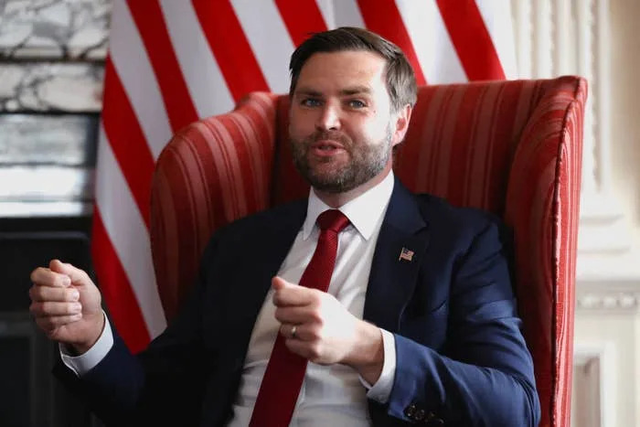 Person in a suit sits in a red-striped chair with a U.S. flag backdrop, gesturing with hands as if speaking