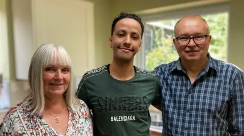 BBC Anita is on the left of the picture. She has shoulder length white hair and is wearing a flowery blouse and a gold necklace. Aziz is in the middle and has short black hair and a green T-shirt that says "Sinners: Baleciaga". Steve is on the right of the picture and he is wearing a checked blue and dark blue buttoned shirt and he has dark glasses. He is balding but has a bit of short hair on his head.