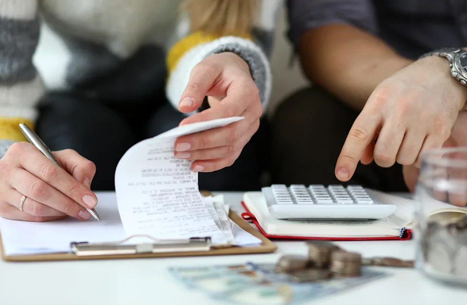 Two people calculating expenses, with one holding a receipt and pen, the other pointing at a calculator. Coins and bills are on the table