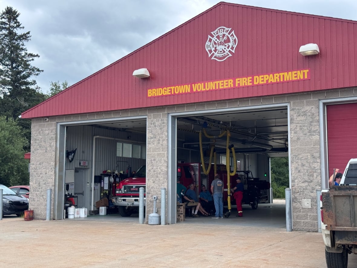 A fire station with a sign that says Bridgetown Volunteer Fire Department has its vehicle bays open with people standing inside.