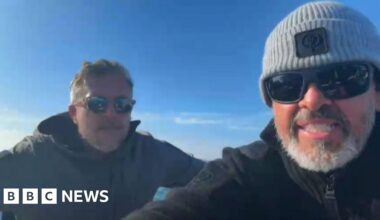 Two men sat next to each other on a boat. Matthew Parker (right) is wearing a grey beanie hat, black coat and black sunglasses. He is sat next to Mike Bates who is wearing a black coat and sunglasses.