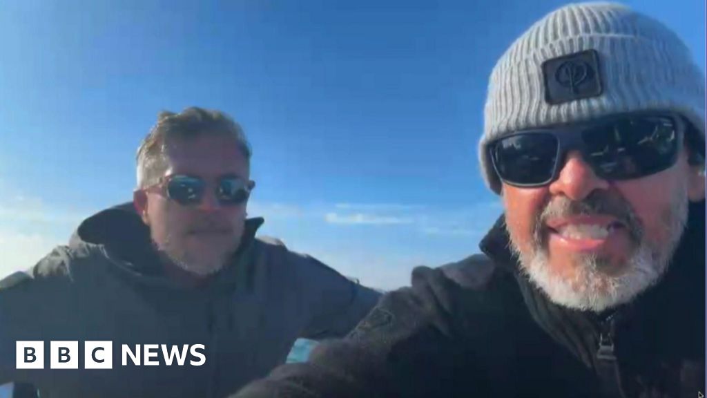 Two men sat next to each other on a boat. Matthew Parker (right) is wearing a grey beanie hat, black coat and black sunglasses. He is sat next to Mike Bates who is wearing a black coat and sunglasses.