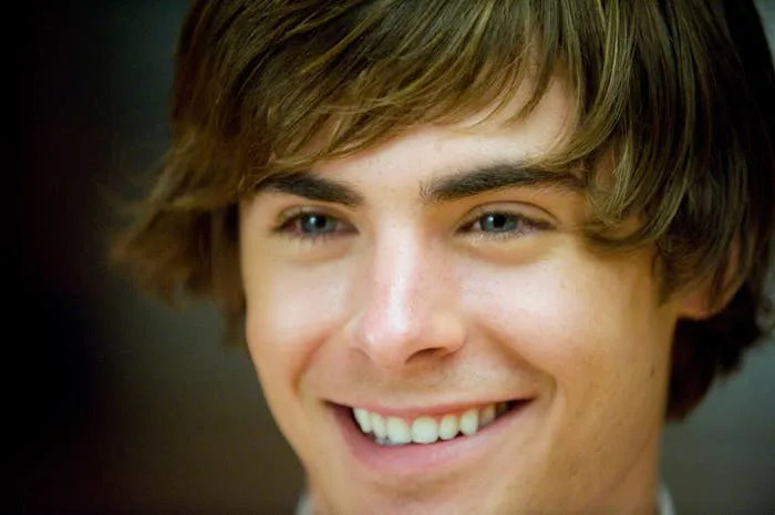 Zac Efron smiling warmly with medium-length brown hair, captured in a close-up shot