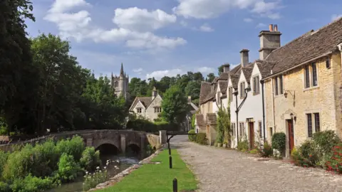 A picturesque village with a row of house, a cobble lane, a river and a stone bridge. In the background, there is a church. 