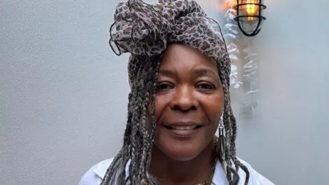 BBC Annetta Stanley is looking directly at the camera. She has long, braided, hair and is wearing a scarf, tied in a bow, on her head. She is wearing a white shirt and a gold coloured necklace.