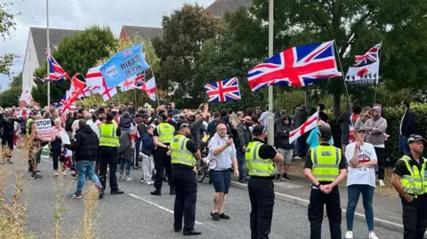 BBC A group of people carrying Union Jacks and England flags in a suburban street. A line of police look on.