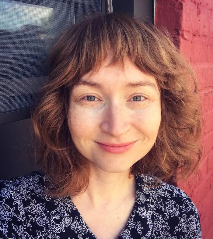 Person with wavy hair smiling, wearing a patterned top, standing in front of a brick wall