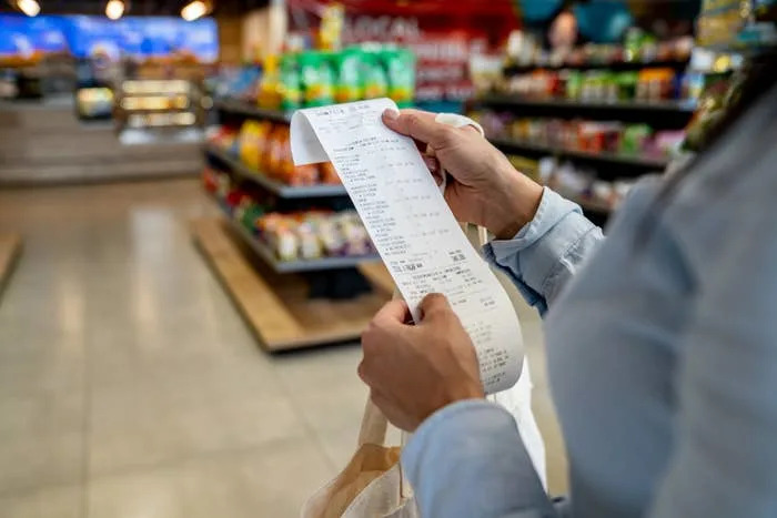Person holding a shopping receipt in a grocery store aisle, reviewing purchases. Shelves stocked with various products are in the background