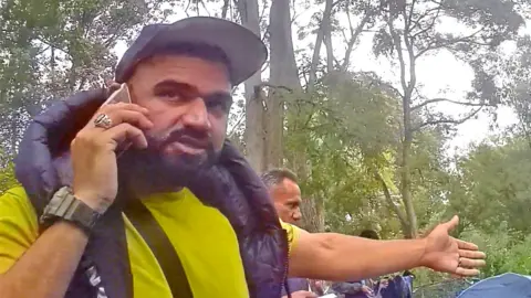 Abdullah the smuggler, in a forest clearing, with people in the background behind him.