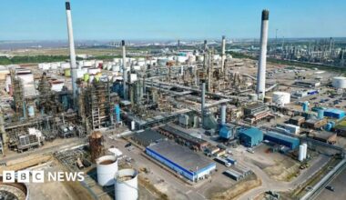 Aerial view of the Lindsey Oil Refinery, a large industrial complex with numerous tall chimneys, large cylindrical storage tanks and a network of interconnected pipelines. Various buildings can be seen throughout the facility, which is surrounded by roads and open land beneath a clear blue sky.