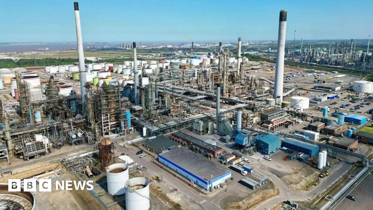 Aerial view of the Lindsey Oil Refinery, a large industrial complex with numerous tall chimneys, large cylindrical storage tanks and a network of interconnected pipelines. Various buildings can be seen throughout the facility, which is surrounded by roads and open land beneath a clear blue sky.