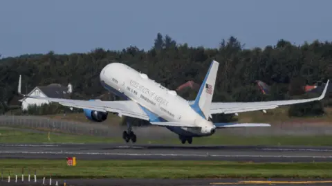 Reuters The Air Force Two plane taking off at Prestwick Airport. The planes wheels are just off the ground. It is mainly white with blue accents and the engines are also covered with a blue metal material. The words 'United States of America' are in a  thin gold font on the side of the plane.