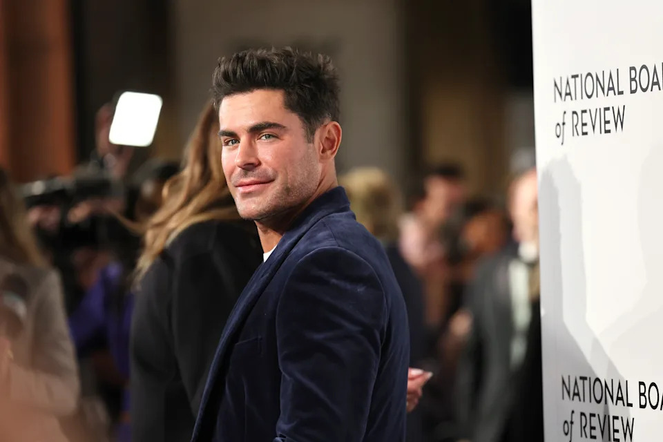 Zac Efron in a velvet jacket on a red carpet at a National Board of Review event, smiling at the camera