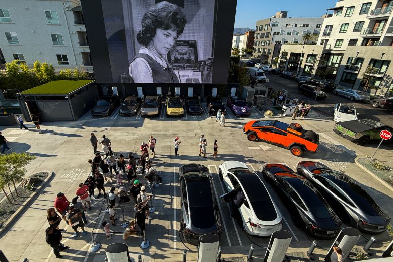 People wait in line as Tesla electric vehicles, including the Cybertruck, park beneath a giant movie screen at the Tesla Diner.