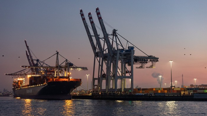 Cranes extend above a container ship at dusk in the Port of Hamburg, Germany