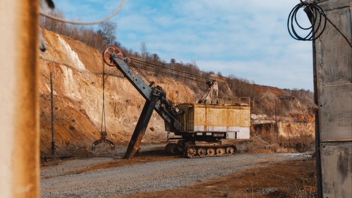 A large, yellow excavator with a claw attachment operates in an open-pit graphite quarry, surrounded by exposed rock walls.