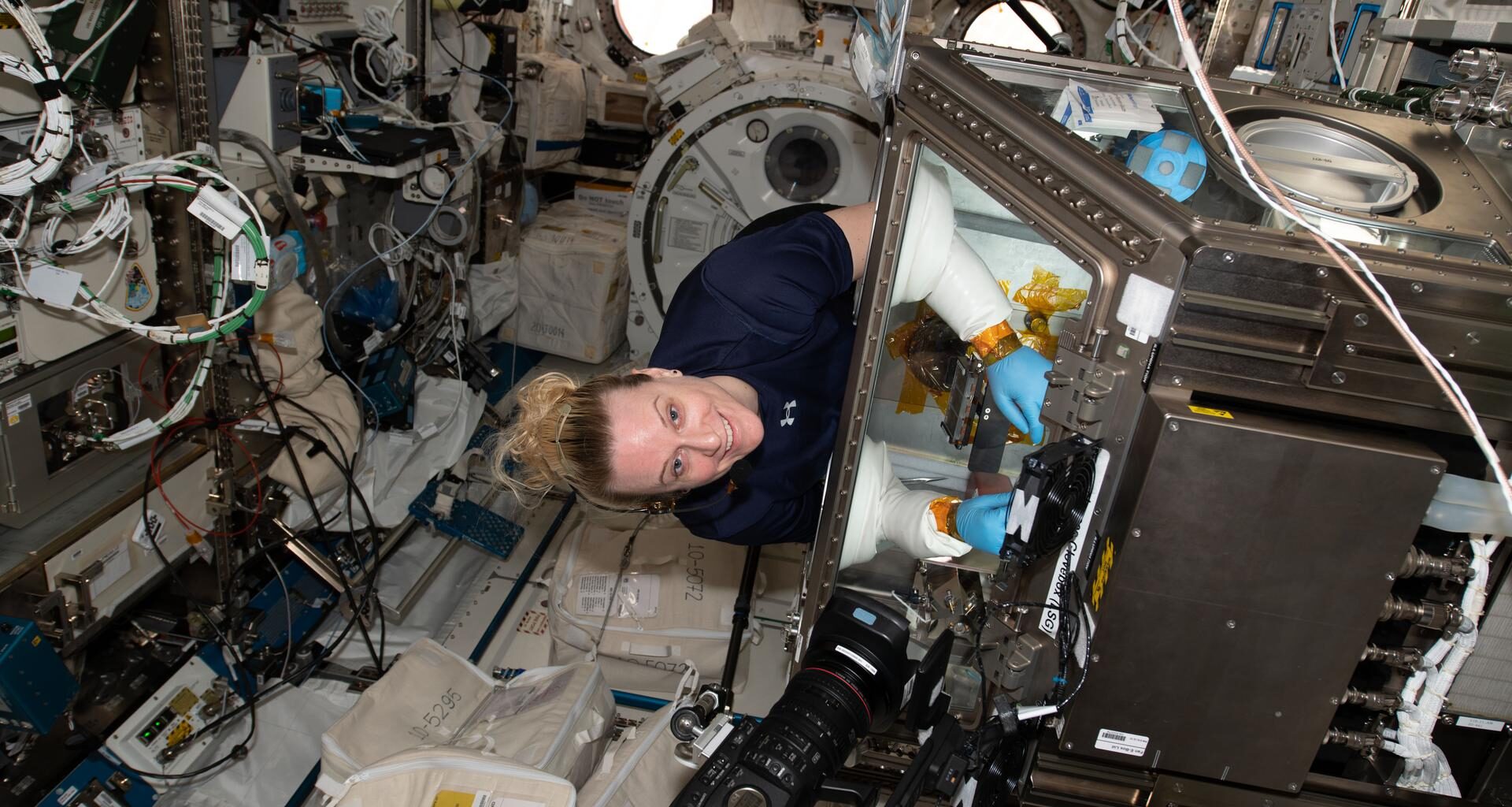 Rubins stands at an angle wearing a navy-blue shirt, smiling while conducting an experiment inside a glovebox. The metallic glovebox is about the size of a washing machine, with a glass cover. Rubins’ arms are inserted through the glass cover, wearing protective blue latex gloves and white protective sleeves. A black camera is pointed towards the glovebox, and the surrounding walls have many wires, hardware, and containers attached.