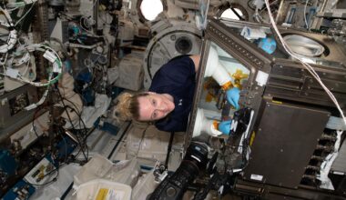 Rubins stands at an angle wearing a navy-blue shirt, smiling while conducting an experiment inside a glovebox. The metallic glovebox is about the size of a washing machine, with a glass cover. Rubins’ arms are inserted through the glass cover, wearing protective blue latex gloves and white protective sleeves. A black camera is pointed towards the glovebox, and the surrounding walls have many wires, hardware, and containers attached.