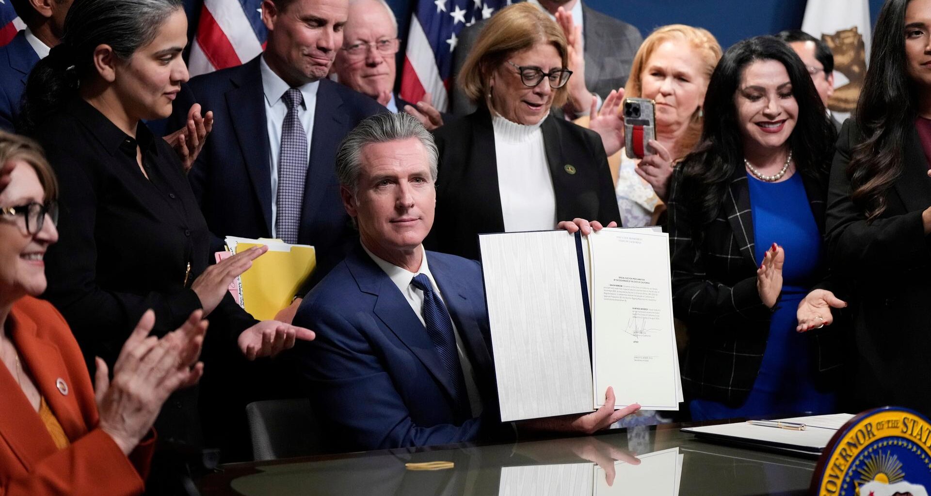 Newsom sitzt an seinem Gouverneurstisch und hält das Dokument in die Kameras. Hinter und neben ihm stehen zahlreiche Parteifreunde.