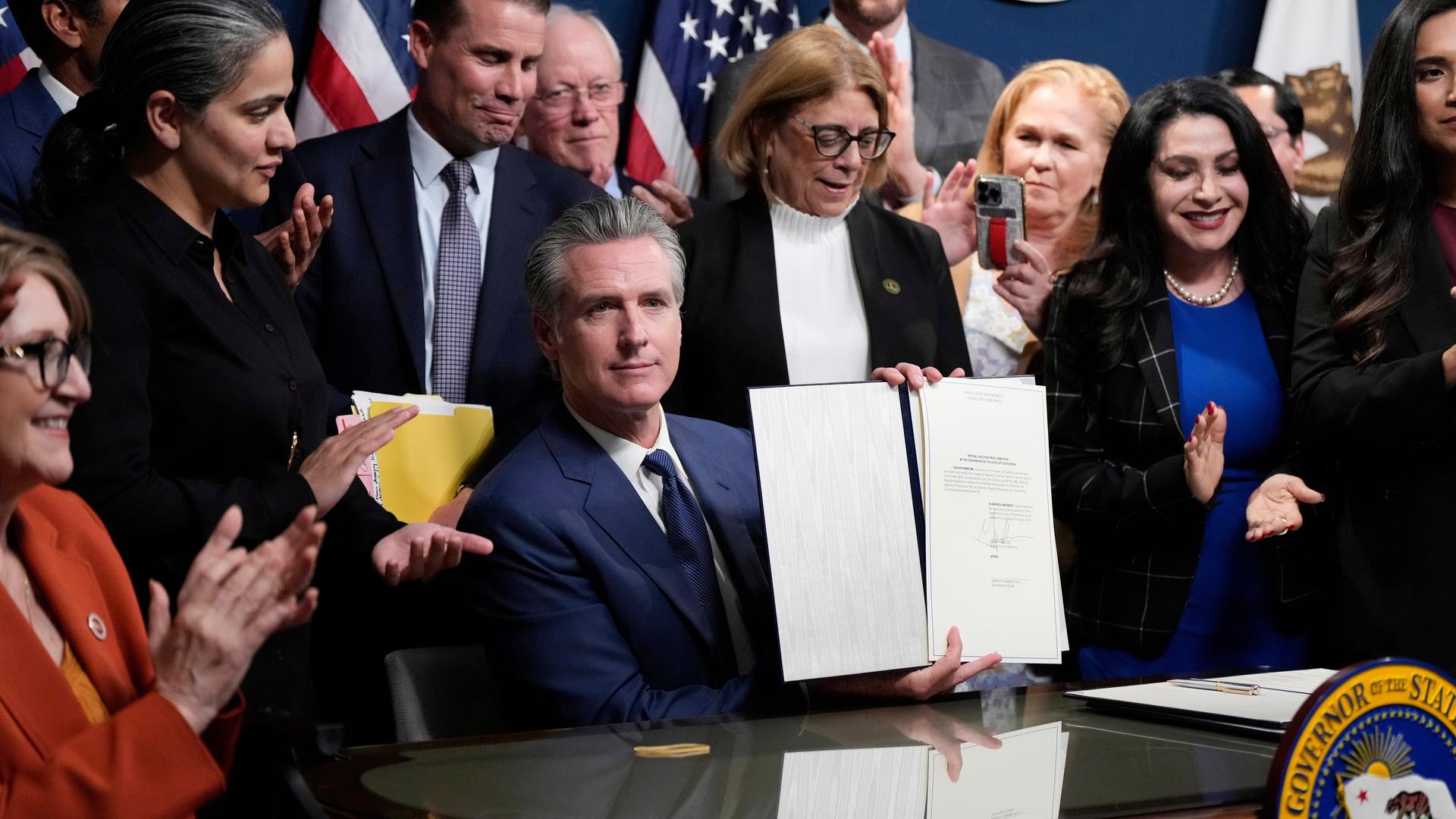 Newsom sitzt an seinem Gouverneurstisch und hält das Dokument in die Kameras. Hinter und neben ihm stehen zahlreiche Parteifreunde. 