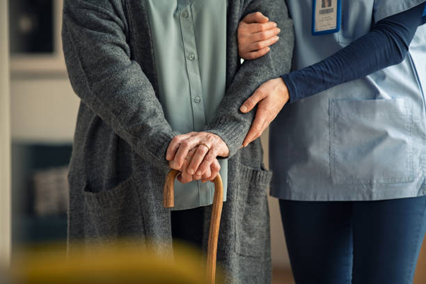 A caregiver supports an elderly person holding a cane, gently guiding them by the arm.