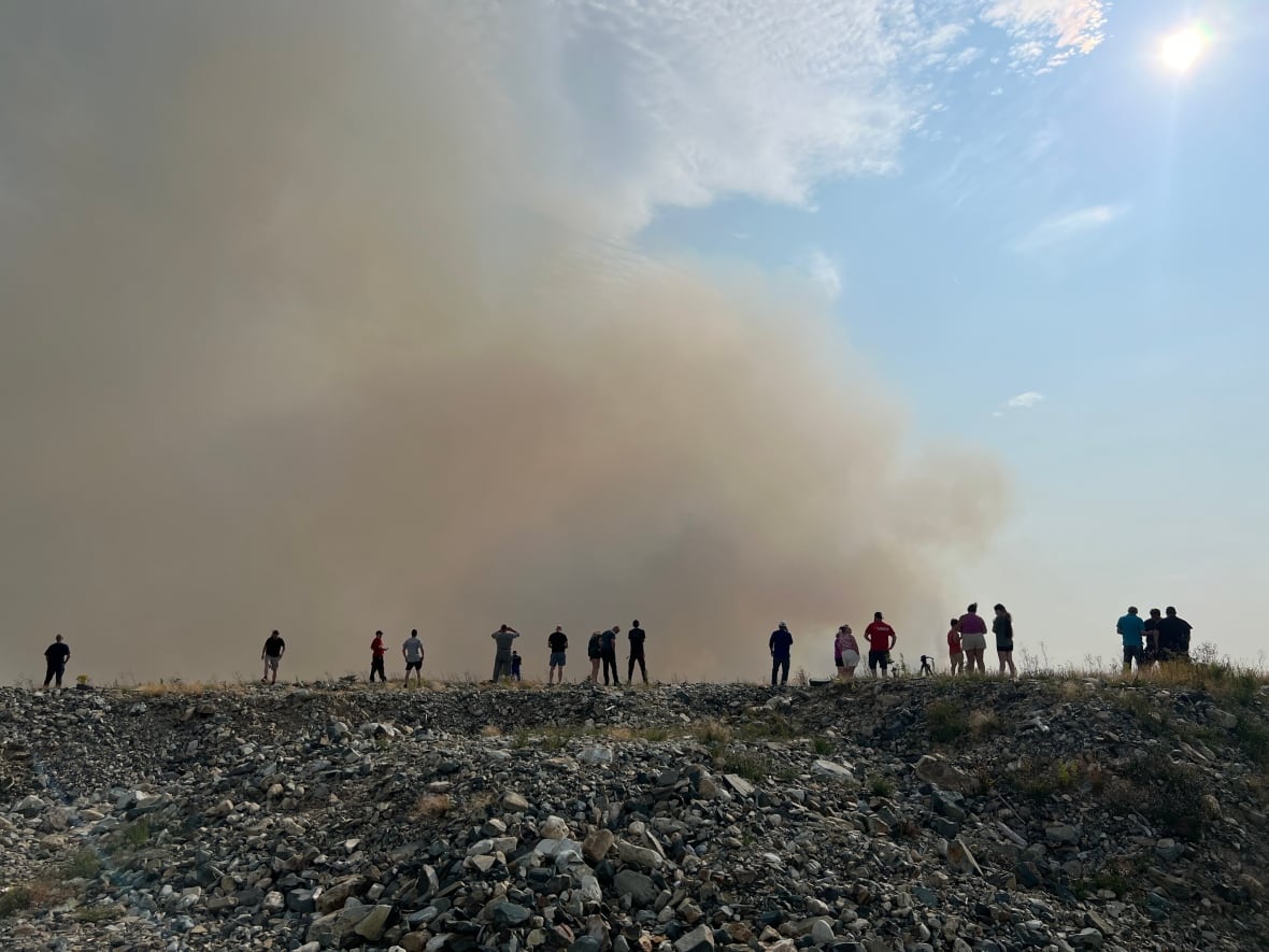 A line of people stand on a hill watching smoke rise beyond it.