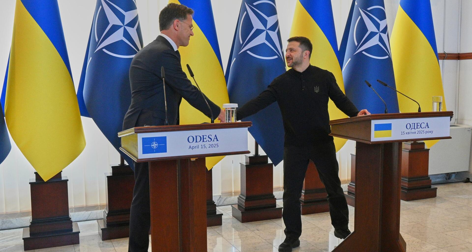 Die beiden stehen an Rednerpulten und schütteln sich die Hand. Dahinter mehrere Flaggen der Nato und der Ukraine.