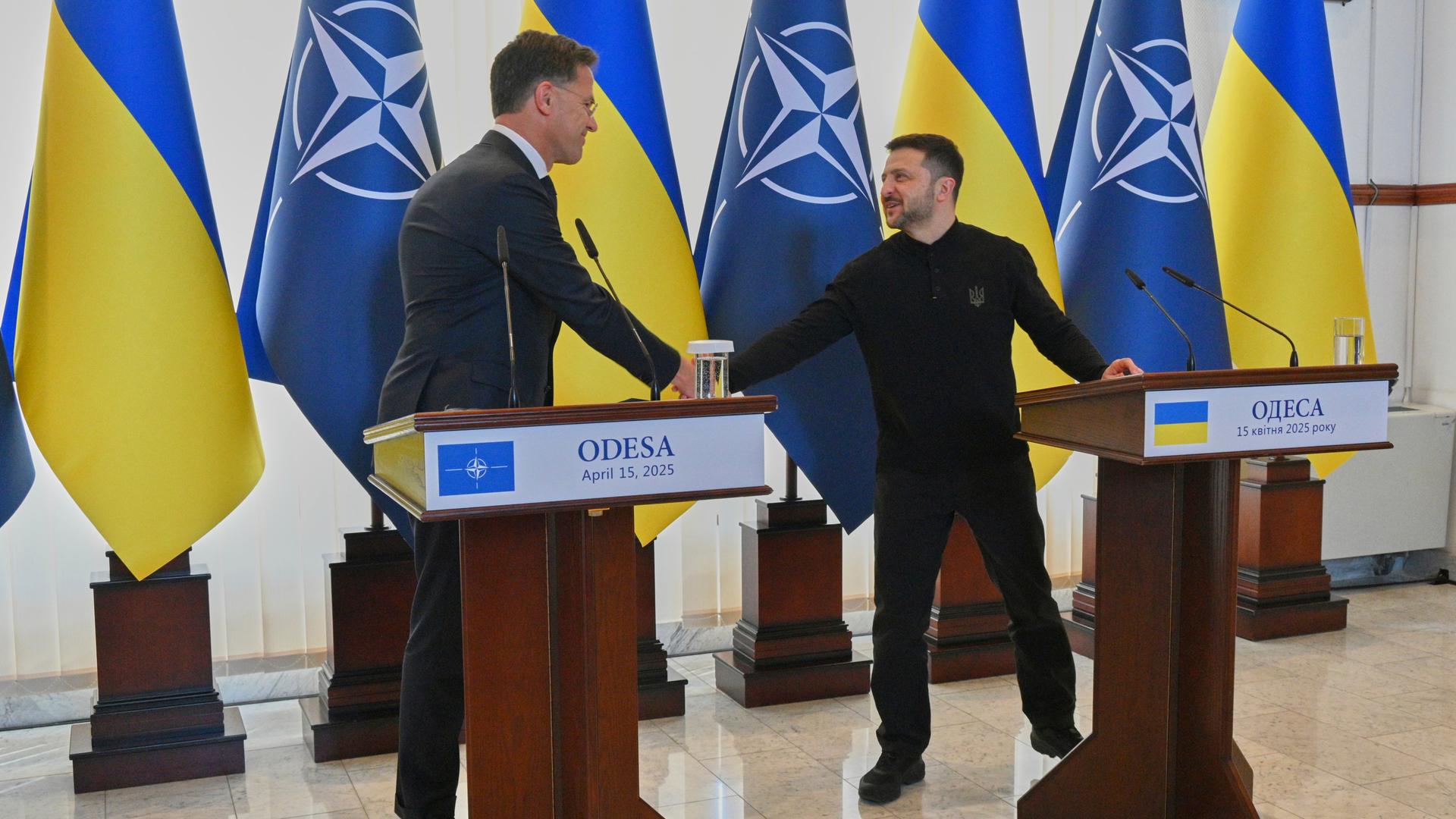 Die beiden stehen an Rednerpulten und schütteln sich die Hand. Dahinter mehrere Flaggen der Nato und der Ukraine. Die beiden stehen an Rednerpulten und schütteln sich die Hand. Dahinter mehrere Flaggen der Nato und der Ukraine.