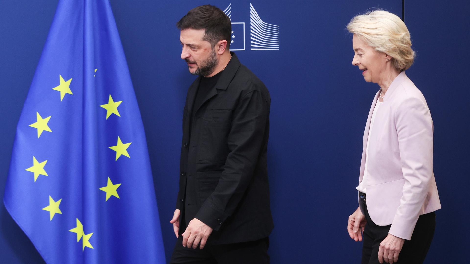 Der ukrainische Präsident Selenskyj und EU-Kommissionspräsidentin von der Leyen stehen vor einem blauen Hintergrund mit einer europäischen Flagge.