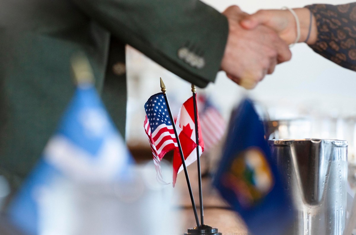 Two people shaking hands near small U.S. and Canada flags on a table, with blurred flags in the foreground.