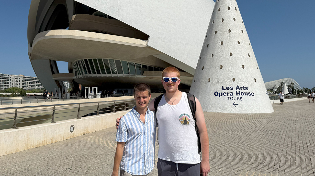 Two young men stand in front of a music hall with a contemporary design.
