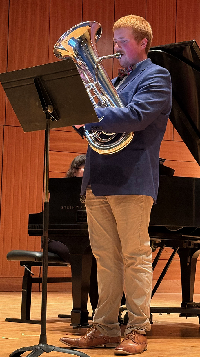 A young man wearing a suit plays a euphonium on a stage in a performance hall.